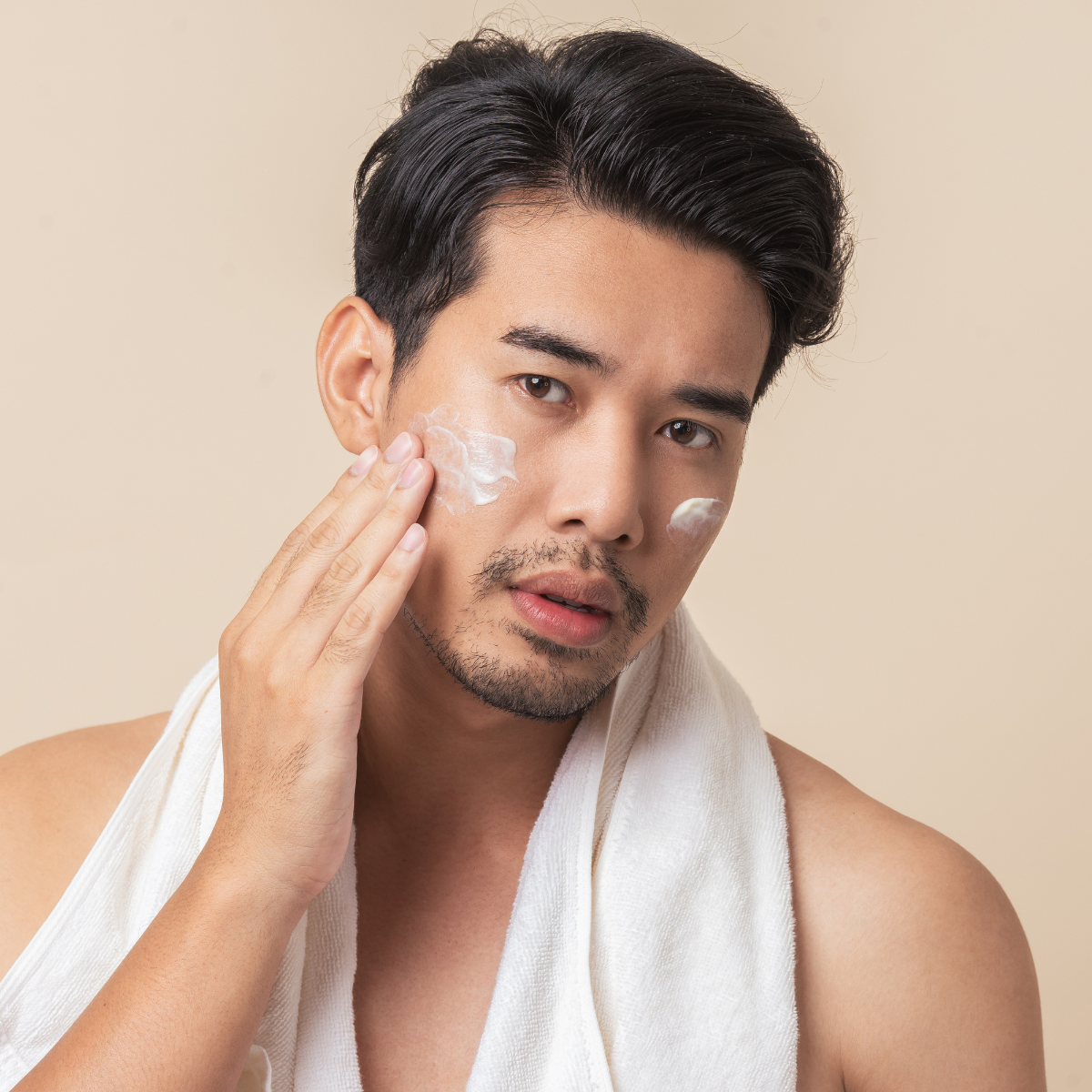 Man applying cream to his face with a towel around his neck against a beige background
