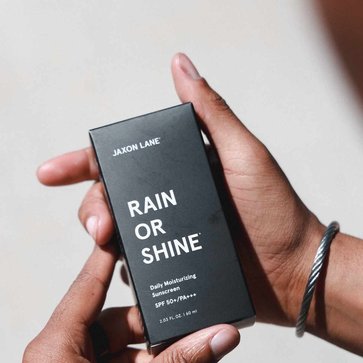 Hand holding a Jaxon Lane 'Rain or Shine' sunscreen box against a neutral background